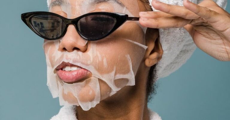 Skincare Routines - Self esteem young African American lady with moisturizing sheet mask and towel on hand wearing trendy sunglasses while standing against blue background in bathrobe after shower