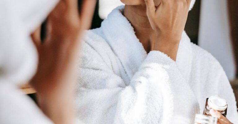 Cleanse - Young black female in white robe and towel on head applying moisturizing cream on face while standing in bathroom